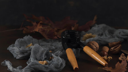 A heavy-duty black and wood nutcracker sits among walnuts and chestnuts on a dark wooden surface. Soft lighting highlights the rustic textures of gauze fabric and dried oak leaves.の写真素材
