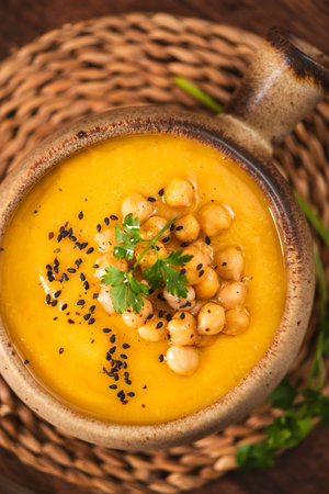 A warm bowl of vibrant butternut squash or pumpkin puree served in a rustic ceramic pot. The dish features textured toppings of whole chickpeas and fresh parsley under soft, natural lighting.の写真素材