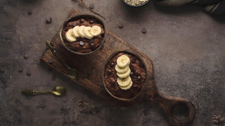 Two bowls of rich chocolate porridge topped with sliced bananas and dark chocolate chips. The overhead shot features moody lighting, textured stone surfaces, and vintage spoons.の写真素材