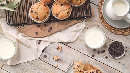 Freshly baked golden muffins with dark chocolate morsels sit on a cooling rack over a wooden board. Soft natural lighting enhances the cozy, rustic kitchen aesthetic with glasses of fresh milk.の写真素材