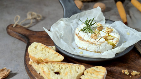 Warm melted Camembert topped with fresh rosemary and honey sits on crumpled parchment paper. The rustic scene features a dark skillet and crusty bread under soft, moody kitchen lighting.の写真素材