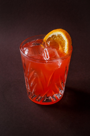 Glass of red alcohol cocktail with ice and slice of orange on elegant dark brown background.の写真素材