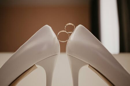 Gold wedding rings between pair of white high heel shoes. Wedding details on the table near mirror.の写真素材