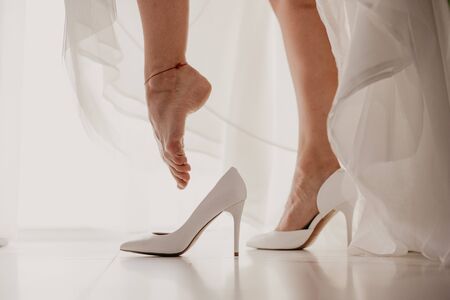 Bride wearing high heel shoe on mirror floor near panoramic window on wedding day. Bride putting her elegant leg into white shoe.の写真素材