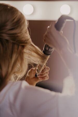 Bride twists the hair with a curling iron at home. Preparation of the bride. Close up. Women doing hairstyle herself and using straightener on beautiful long curl hair. Beauty and haircare concept.の写真素材
