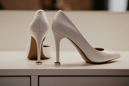 2 Two Gold wedding rings under pair of white high heel shoes. Wedding details on white table near mirror.の写真素材