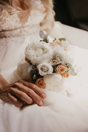 Wedding bouquet in hands of bride. Beautiful bride sitting and holding wedding bouquet with beautiful roses and pions. In white dress and nice monicure. Close up.の写真素材