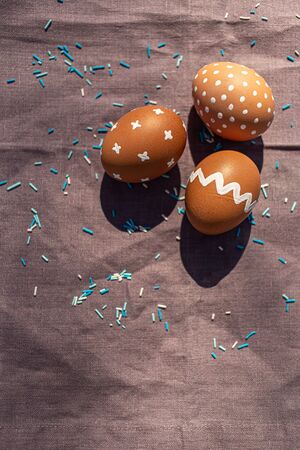 Painted 3 three brown eggs with various white patterns for Easter holiday on cotton purple tablecloth with sunlights and shadows. Decoration blue candies. Top view. Eastern European Orthodox cultures.の写真素材
