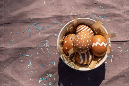 Painted brown eggs with various white patterns for Easter holiday in bowl on cotton purple...