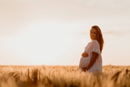 Young beautiful pregnant woman looking to the side and touching her belly in the wheat orange field on a sunny summer day. Nature in the country. Miracle expectation. Sunset on isolation. Copy Spaceの写真素材