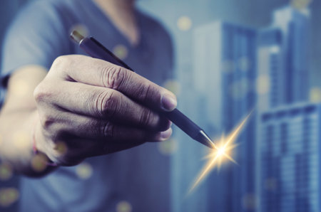 human hand holding a pen with a sparkling light at the nib. The background consists of blue skyscrapers slightly out of focus. The image suggests creativity and the power of writing.の写真素材