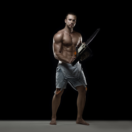 Young muscular man with chainsaw posing on black backgroundの写真素材