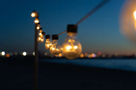 Close up of the glowing light bulbs at the beach cafe terrace. Sun is setting in the blurry abstract background.の写真素材