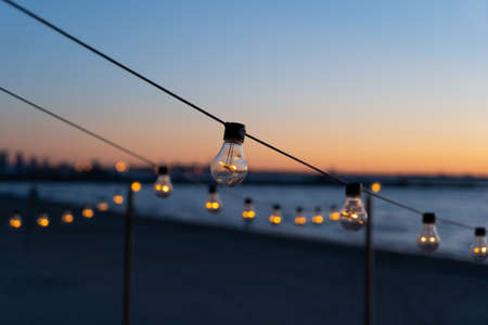 Close up of the glowing light bulbs at the beach cafe terrace. Sun is setting in the blurry abstract background.の写真素材