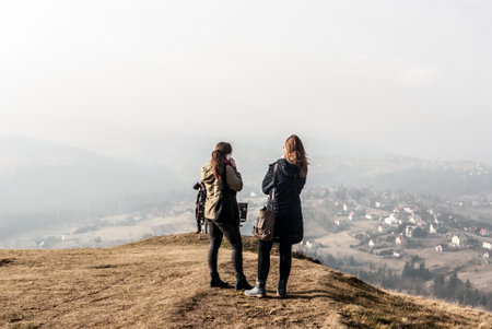 two young nice polish girls (one of them phoning) with long brown hair on Ochodzita hill summit with Koniakow village bellow in late autumn Silesian Beskids mountains in Polandのeditorial素材