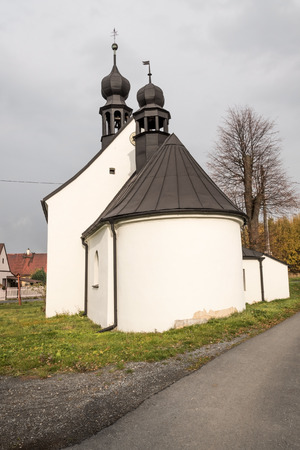 village scenery white white coloured chapel, houses on the background, road, grass and colorful trees on Krizov settlement near Spvinec castle in Czech republicの写真素材