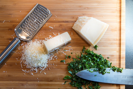 grated parmesan cheese, chopped basil, knife and metal grater on wooden plateの写真素材