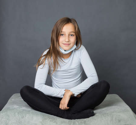 Portrait of a young girl sitting on pillow with medical mask on her chin on grey background with space for textの写真素材
