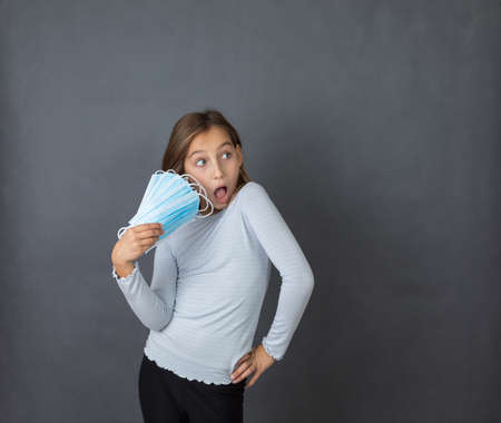 Portrait of a young scared girl with open mouth with fan made of medical masks in her hand on grey background.の写真素材