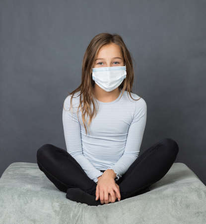 Portrait of a young teen girl in a medical mask sitting on the ground with grey background.の写真素材