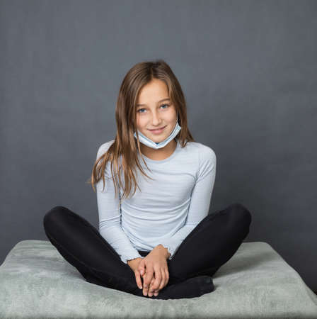 Portrait of a young teen girl smiling in a medical mask sitting on the ground with grey background.の写真素材