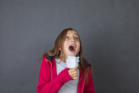 Portrait of a young girl trying to breath with open mouth in red sweater playing with medical mask on grey background.の写真素材
