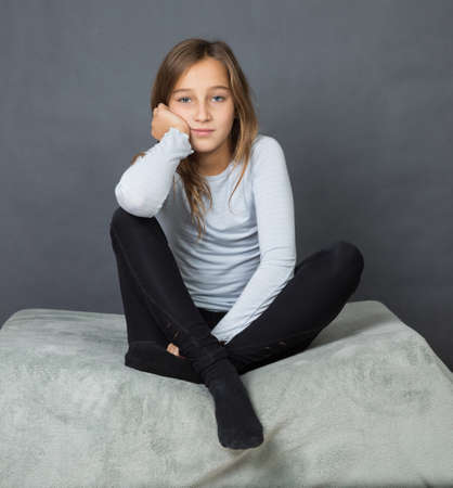 Portrait of a young girl sitting on pillow on grey background with hand on her faceの写真素材