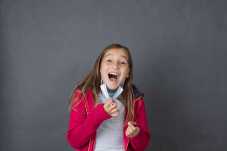 Portrait of a young happy girl trying to breath with open mouth in red sweater playing with medical mask on grey background.の写真素材