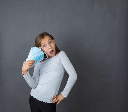 Portrait of a young scared girl looking into camera with open mouth with fan made of medical masks in her hand on grey background.の写真素材