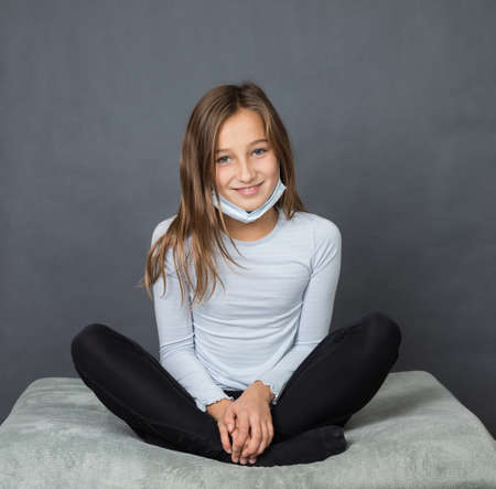 Portrait of a young smiling girl sitting on pillow with medical mask on her chin on grey background with space for textの写真素材
