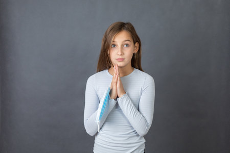 Portrait of a young praying girl playing with medical mask on grey background.の写真素材