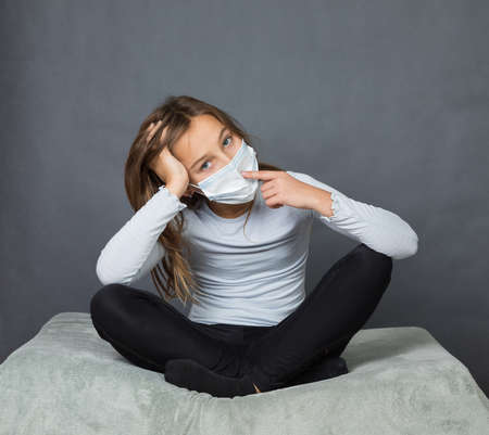 Portrait of a young teen girl in a medical mask with finger on her nose sitting on the ground with grey background.の写真素材
