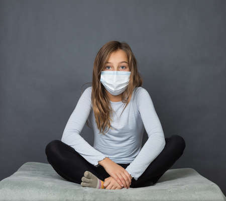 Portrait of a young teen girl in a medical mask sitting on the ground with grey background.の写真素材