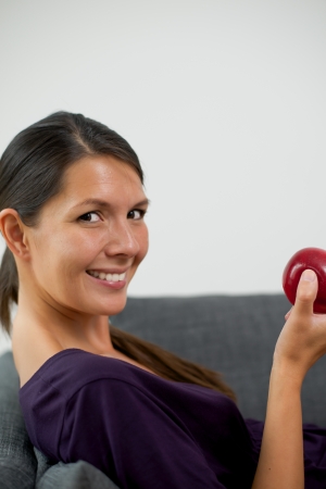 Pretty young woman holding a fresh crisp red apple in her hand while relaxing on a sofa at home giving the camera a lovely friendly smileの写真素材