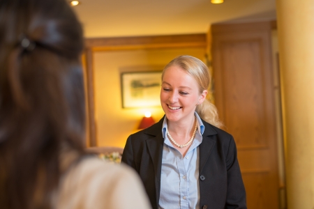 Beautiful stylish blond hotel receptionist standing behind the service desk in a hotel lobby looking at the camera with a friendly smileの写真素材