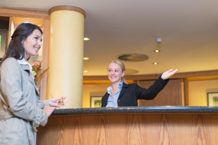 Low angle view of a beautiful friendly smiling receptionist behind the service desk in a hotel lobby helping an attractive female guest indicating with her hand the way to her accomodationの写真素材
