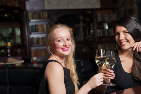 Two beautiful elegant women friends in stylish simple black cocktail dresses toasting each other with glasses of chilled champagne as they celebrate on a night out togetherの写真素材