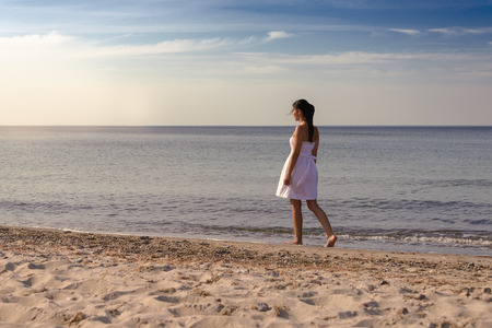 Meditative beautiful young woman standing sideways on a tropical beach in a fresh white summer dress looking down at the ground with a serene expression against an ocean backdrop with copy spaceの写真素材