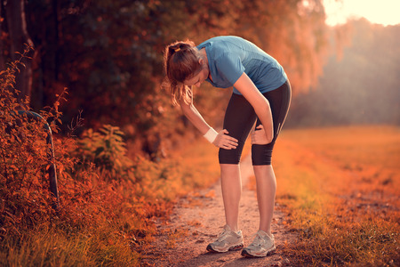 Young athletic woman taking a break from training standing resting her hands on her knees on a rural track through lush farmland in a health and fitness conceptの写真素材