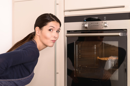 Pretty Young Woman Wearing Apron Waiting the Baked cake Near The Oven While looking at the Camera.の写真素材