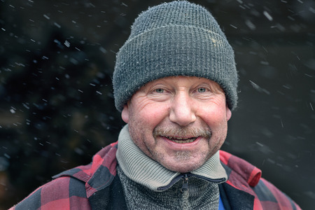 Happy man with a mustache in a knitted winter cap and warm winter clothing looking at the camera with a charismatic friendly smile as he stands outdoors in snowの写真素材