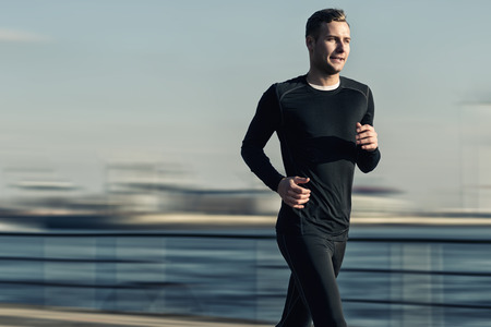 Athletic Handsome Man in Black Workout Clothing Running at the River Bridge Early in the Morning.