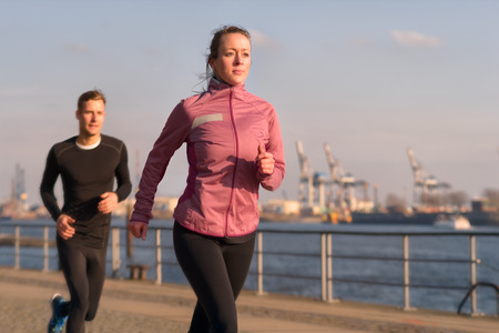 Fit active young couple running on a seafront promenade with the athletic woman leading the way approaching the camera in a healthy lifestyle conceptの写真素材