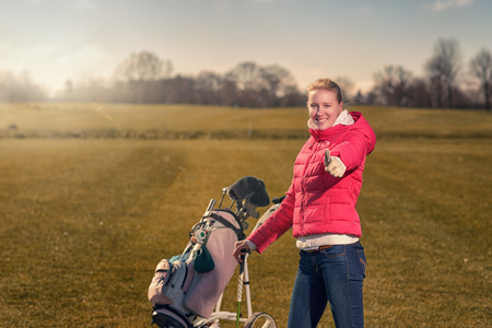 Smiling happy attractive young female golfer giving a thumbs up of success and approval as she pushes her golf cart across the greenの写真素材