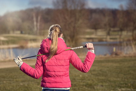 Rear View of a Blond Female Golfer in Dark Pink Jacket Holding a Golf Club on her Shoulder While Looking Afar, Captured in Half Body Shot.の写真素材