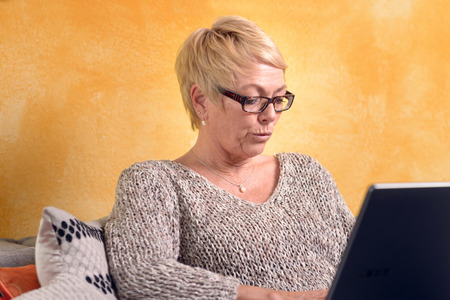 Serious Middle Age Woman with Short Blond Hair Sitting on Sofa While Using her Laptop Computer.の写真素材