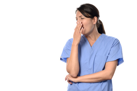 Exhausted attractive female doctor or nurse in blue scrubs standing yawning with her hand to her mouth of a long tiring shift, isolated on whiteの写真素材