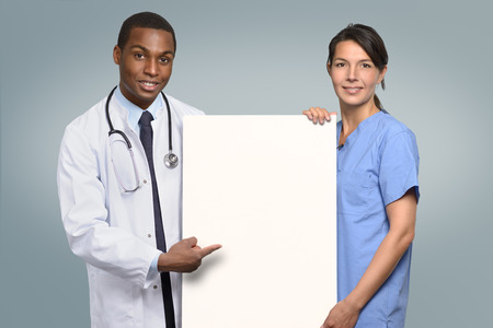 Multiethnic medical team of an African doctor in a lab coat and stethoscope and a nurse in scrubs holding a blank white sign with copyspce for your text pointing to it with happy friendly smilesの写真素材