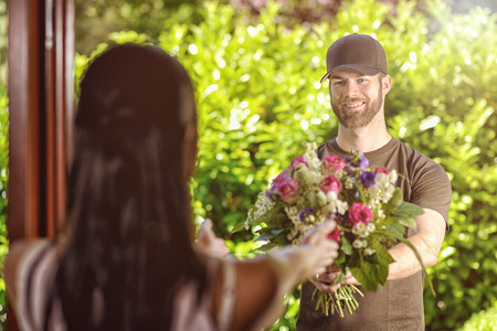 Smiling bearded 20s man wearing brown cap and brown t-shirt delivers flowers to door of young brunette female. Over the shoulder rear view of brunette.の写真素材