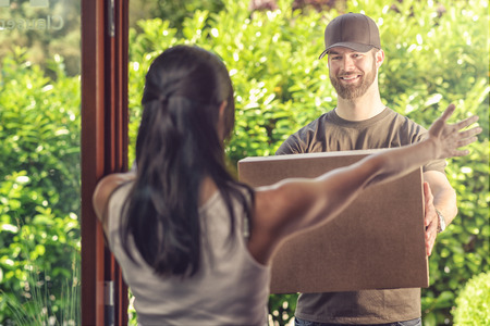Woman taking delivery of a large parcel holding out her arms to the smiling friendly deliveryman as he hands it over, view from behind over her shoulderの写真素材